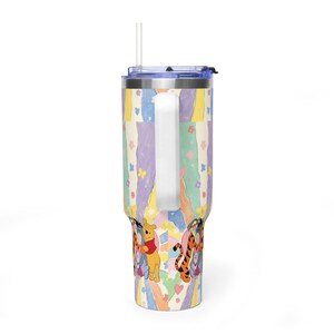 Puede incluir: Un vaso de acero inoxidable con asa, tapa transparente y pajita. El exterior presenta un patr&oacute;n de rayas verticales en colores pastel con peque&ntilde;as flores, mariposas y corazones, junto con ilustraciones de Winnie the Pooh, Igor y Tigger.