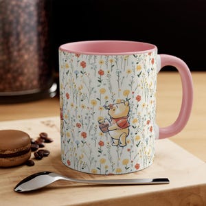 May include: A ceramic mug with a pink interior and handle, featuring a floral pattern and a Winnie the Pooh illustration. The mug has a white background with colorful flowers and a cartoon bear holding a honey pot.