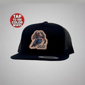 May include: Black trucker hat with a mesh back and a flat brim. The hat features a patch with a dog holding a bird, with the text "GAME BIRD RETRIEVAL". A red circle with the text "TAP TO SEE COLOR WAYS" is in the upper left.