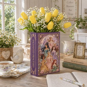 May include: A purple rectangular vase with the words "ENCHANTED PRINCESSES" on the side, filled with yellow tulips and white baby's breath. The vase features illustrations of princesses. A teacup, books, and a framed picture are nearby.