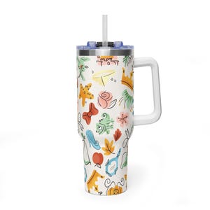 May include: A white tumbler with a handle and a straw, covered in a colorful pattern of cartoon illustrations. The design includes a crown, a rose, a bow, a fish, a shoe, and a pumpkin. The tumbler has a blue lid and a white handle.