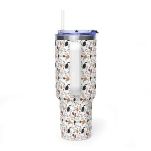 Op de afbeelding: Een witte beker met een handvat en een deksel, versierd met een herhalend patroon van zwarte, bruine en oranje kattenillustraties. De beker heeft een doorzichtig rietje en een blauw deksel. Het ontwerp is een speels thema.