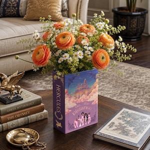 Puede incluir: Un arreglo floral de ranúnculos naranjas y margaritas blancas en un jarrón con forma de libro. El jarrón con forma de libro presenta el texto "Hercules" y "Go the Distance" con una ilustración de castillo.