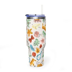 May include: A white tumbler with a blue lid and straw, covered in colorful cartoon illustrations. The design includes a crown, rose, bow, and other whimsical elements. The tumbler is designed to hold approximately 30 ounces.