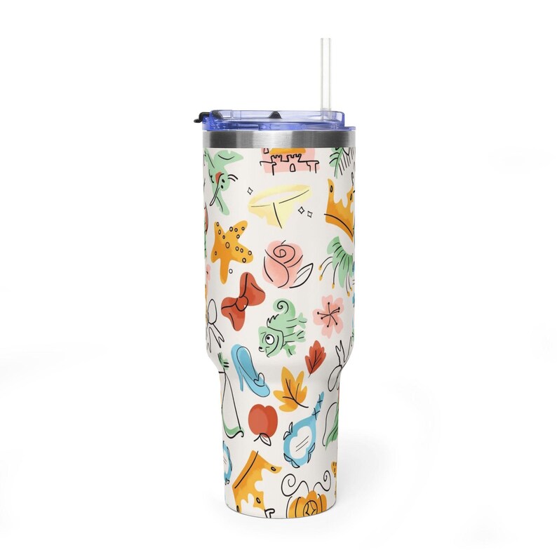 May include: A white tumbler with a blue lid and straw, covered in colorful cartoon illustrations. The design includes a crown, rose, bow, and other whimsical elements. The tumbler is designed to hold approximately 30 ounces.