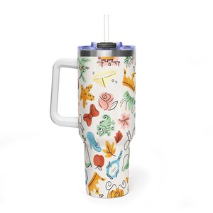 May include: A white travel mug with a handle and a straw, decorated with colorful cartoon illustrations. The design includes a crown, a bow, a shoe, and other whimsical elements. The mug is designed to hold a beverage.
