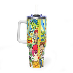 May include: A large, insulated travel mug with a white handle and straw. The mug features a vibrant design with characters from the film "Toy Story", including Woody, Buzz Lightyear, and the Little Green Men. The background has yellow stripes and blue stars.