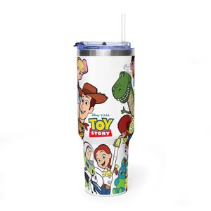 May include: A white tumbler featuring characters from the Disney Pixar movie Toy Story. The tumbler has a blue lid and a clear straw. The Toy Story logo is visible, along with images of Woody, Buzz Lightyear, and other characters.
