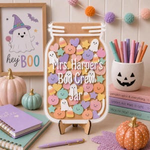 Puede incluir: Un letrero de acrílico transparente con forma de frasco con un fondo color canela lleno de recortes pastel de fantasmas, calabazas, corazones, estrellas y dulces de maíz. El texto "Mrs. Harper's Boo Crew Jar" está impreso en blanco al centro.