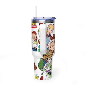 May include: White tumbler featuring characters from the animated film "Toy Story." The tumbler has a handle, a blue lid, and a straw. The design includes Woody, Buzz Lightyear, Jessie, and other characters from the movie.