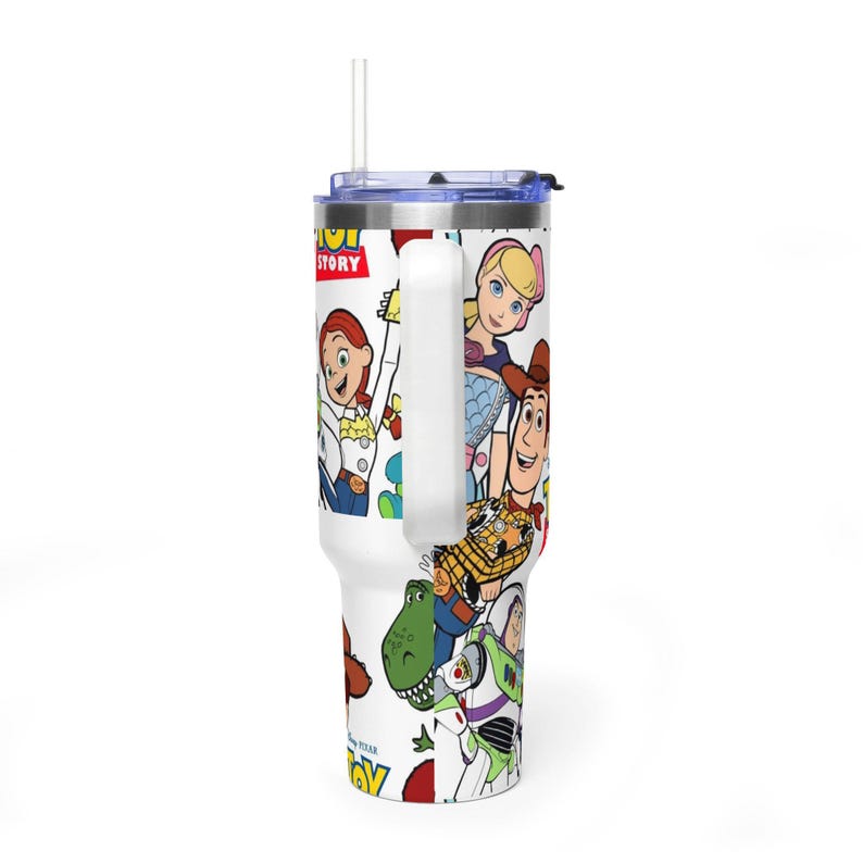 May include: White tumbler featuring characters from the animated film "Toy Story." The tumbler has a handle, a blue lid, and a straw. The design includes Woody, Buzz Lightyear, Jessie, and other characters from the movie.