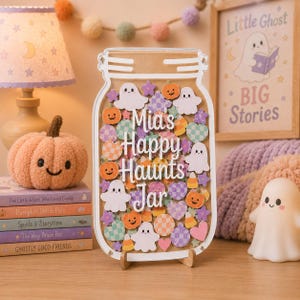 Puede incluir: Un tablero de exhibición de acrílico transparente con forma de frasco que presenta el texto blanco Mia's Happy Haunts Jar. El interior está lleno de formas de madera planas y coloridas, incluyendo fantasmas blancos y calabazas naranjas.