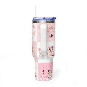 May include: A white and pink tumbler with a Minnie Mouse design. The tumbler has a handle, a straw, and a lid. The design includes Minnie Mouse, trees, and the words "Hello Minnie".