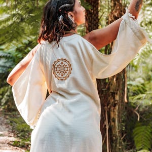 May include: Cream-colored, flowing kimono-style top with wide sleeves and a circular, embroidered design on the back. The top is worn outdoors in a forest setting, with the person's arm raised.