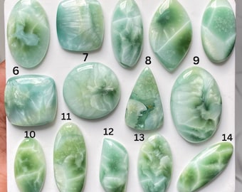 May include: A collection of fourteen polished green gemstones arranged on a white board, numbered one through fourteen. Each stone features unique swirling patterns in shades of light and dark green, with varied shapes including ovals, rectangles, and triangles.