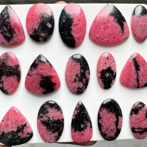 May include: A collection of polished rhodonite cabochons in various shapes including ovals, triangles, and teardrops. Each stone features a distinct pattern of vibrant pink and deep black mineral inclusions with a smooth, reflective surface finish.