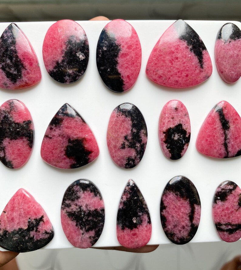 May include: A collection of polished rhodonite cabochons in various shapes including ovals, triangles, and teardrops. Each stone features a distinct pattern of vibrant pink and deep black mineral inclusions with a smooth, reflective surface finish.