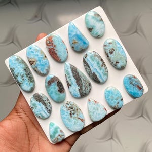 May include: A collection of fourteen polished Larimar cabochons arranged on a white square display board. Each stone features a unique pattern of sky blue, turquoise, and white swirls with dark brown or grey mineral inclusions in oval and teardrop shapes.