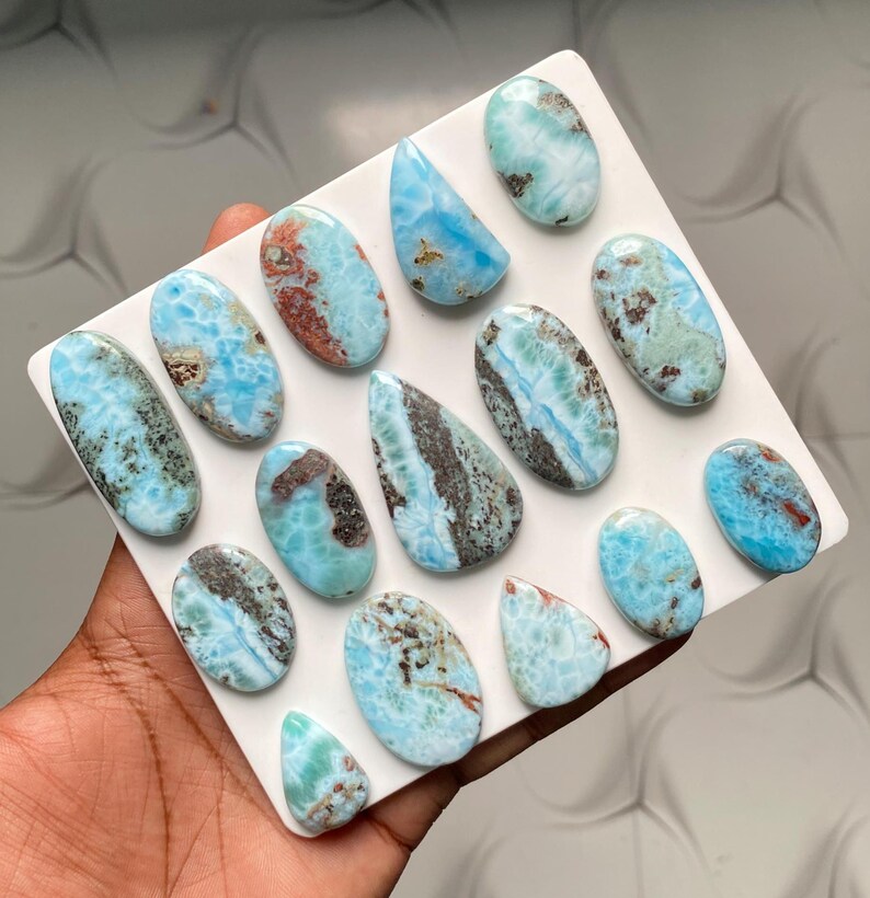 May include: A collection of fourteen polished Larimar cabochons arranged on a white square display board. Each stone features a unique pattern of sky blue, turquoise, and white swirls with dark brown or grey mineral inclusions in oval and teardrop shapes.