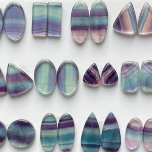 Pu&ograve; includere: Una collezione di cabochon in fluorite lucida in varie forme, tra cui ovali, rettangoli, triangoli e gocce. Ogni pietra presenta distinte bande orizzontali in tonalit&agrave; traslucide di viola, verde acqua, verde e rosa tenue.