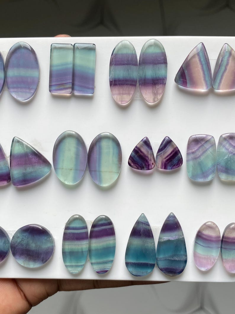 Pu&ograve; includere: Una collezione di cabochon in fluorite lucida in varie forme, tra cui ovali, rettangoli, triangoli e gocce. Ogni pietra presenta distinte bande orizzontali in tonalit&agrave; traslucide di viola, verde acqua, verde e rosa tenue.