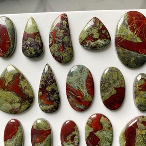 May include: A collection of polished cabochons featuring a mottled pattern of deep red and olive green. These stones are arranged in rows and vary in shape, including oval, teardrop, and irregular forms, all displaying a smooth, glossy finish.
