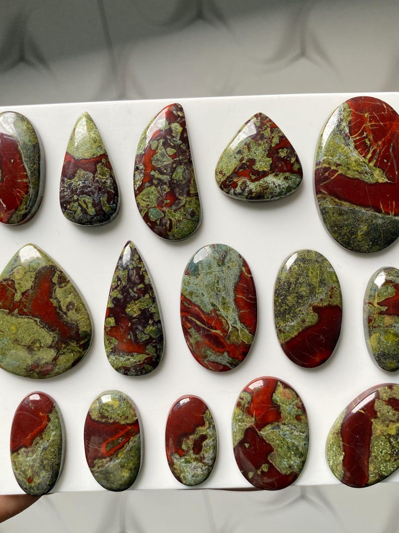 May include: A collection of polished cabochons featuring a mottled pattern of deep red and olive green. These stones are arranged in rows and vary in shape, including oval, teardrop, and irregular forms, all displaying a smooth, glossy finish.