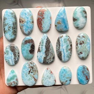May include: A collection of fourteen polished larimar gemstones arranged on a white rectangular display board. The stones feature various shades of light blue, white, and brown, with unique marbled patterns, smooth surfaces, and oval or teardrop shapes.
