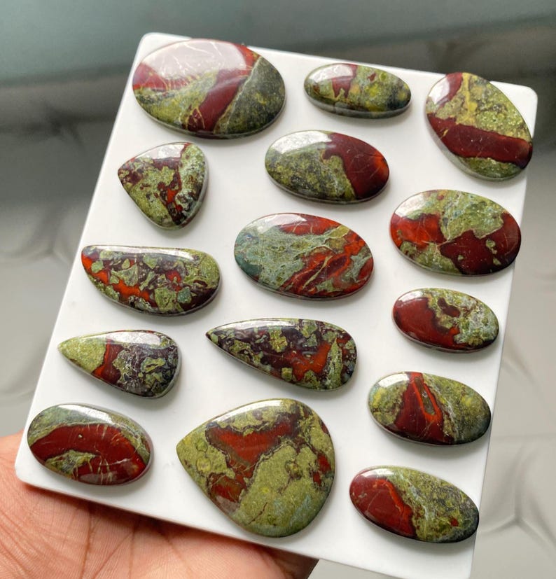 May include: A collection of fifteen polished cabochons featuring a mottled pattern of deep red and olive green stone. These smooth, rounded, and teardrop-shaped gemstones are arranged on a white display board, showcasing varied natural color distributions.