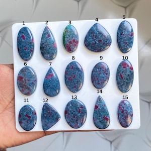 May include: A collection of fifteen polished cabochon gemstones arranged on a white display board, each numbered from 1 to 15. The stones are primarily blue with embedded red inclusions, featuring various shapes including oval, teardrop, and triangular.