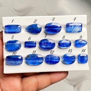 Könnte beinhalten: Eine Sammlung von vierzehn facettierten blauen Kyanit-Edelsteinen, die auf einer weißen Präsentationskarte angeordnet sind. Jeder Stein ist von eins bis vierzehn nummeriert und weist verschiedene Formen wie oval, rechteckig und tropfenförmig auf.