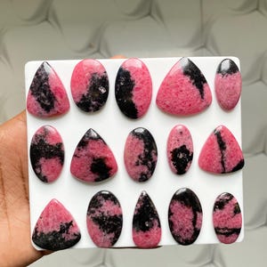 May include: A collection of fourteen polished rhodonite cabochons arranged on a white square display board. Each stone features a distinct pattern of vibrant pink and deep black mineral inclusions, shaped into various ovals, triangles, and teardrops.