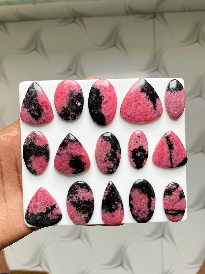 May include: A collection of fourteen polished rhodonite cabochons arranged on a white square display board. Each stone features a distinct pattern of vibrant pink and deep black mineral inclusions, shaped into various ovals, triangles, and teardrops.
