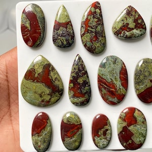 May include: A collection of polished, multi-shaped cabochons featuring a marbled pattern of deep red and olive green. These smooth, rounded stones are arranged on a white surface, displaying varied organic textures and glossy, reflective finishes.
