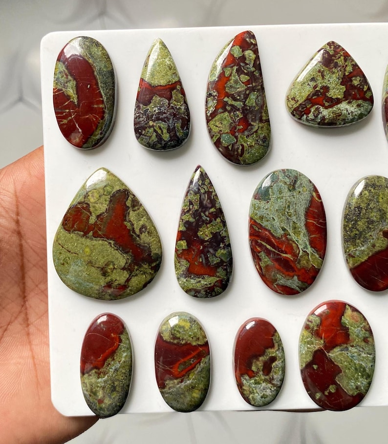May include: A collection of polished, multi-shaped cabochons featuring a marbled pattern of deep red and olive green. These smooth, rounded stones are arranged on a white surface, displaying varied organic textures and glossy, reflective finishes.