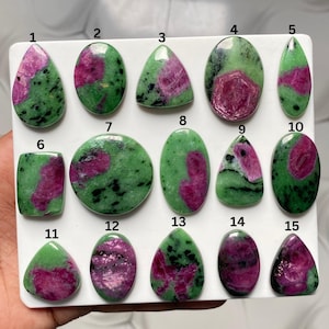 May include: A collection of fifteen polished ruby in zoisite cabochons arranged on a white display board. Each stone features a unique pattern of vibrant green zoisite with inclusions of deep pink ruby, numbered one through fifteen in black text.