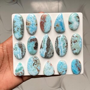 May include: A collection of fourteen polished Larimar gemstones arranged on a white square display board. The stones feature various shapes including ovals and teardrops, displaying marbled patterns of sky blue, white, and dark brown inclusions.