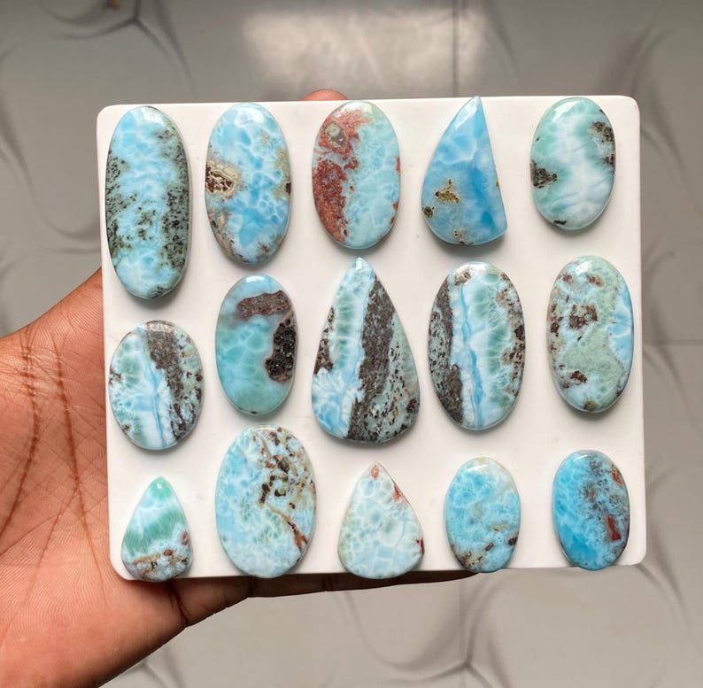May include: A collection of fourteen polished Larimar gemstones arranged on a white square display board. The stones feature various shapes including ovals and teardrops, displaying marbled patterns of sky blue, white, and dark brown inclusions.