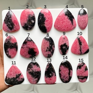 May include: A collection of fifteen polished rhodonite cabochons arranged in three rows on a white display board. Each stone features a unique pattern of vibrant pink and deep black mineral inclusions, numbered one through fifteen for identification.