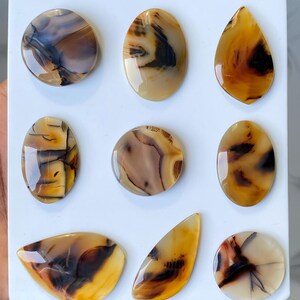 May include: Nine polished agate gemstones in various shapes, including oval, teardrop, and round. The stones display a mix of brown, tan, and black patterns, with a glossy finish. They are arranged on a white surface.
