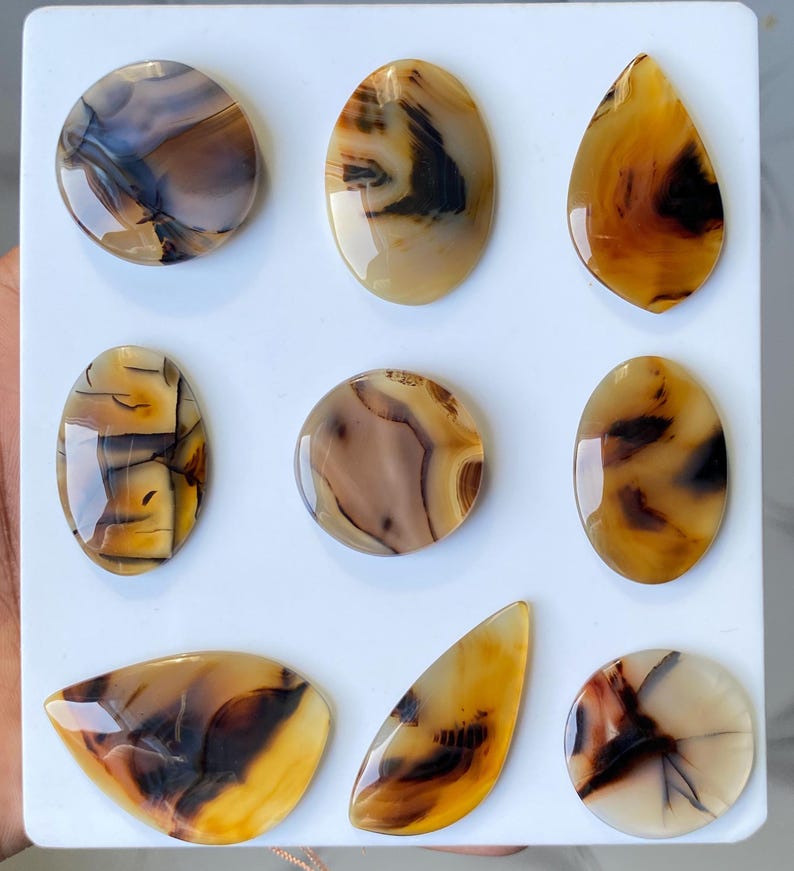 May include: Nine polished agate gemstones in various shapes, including oval, teardrop, and round. The stones display a mix of brown, tan, and black patterns, with a glossy finish. They are arranged on a white surface.