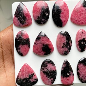 May include: A collection of polished rhodonite cabochons arranged on a white surface. Each stone features a distinct pattern of vibrant pink and deep black mineral inclusions, with various shapes including ovals, triangles, and teardrops.