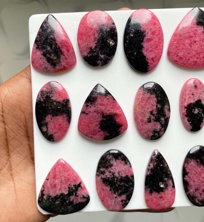 May include: A collection of polished rhodonite cabochons arranged on a white surface. Each stone features a distinct pattern of vibrant pink and deep black mineral inclusions, with various shapes including ovals, triangles, and teardrops.