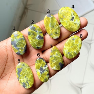 Pode incluir: Uma coleção de oito pedras preciosas ovais e em forma de gota. As pedras apresentam um padrão mosqueado de amarelo-esverdeado e cinza, com variações de cor e veios. As pedras preciosas são seguradas em uma mão.