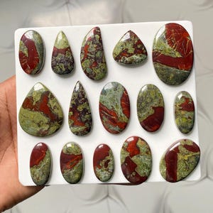 May include: A collection of fourteen polished cabochons arranged on a white rectangular display board. Each stone features a unique pattern of deep red and olive green swirls, with smooth, rounded edges in various oval, teardrop, and irregular shapes.