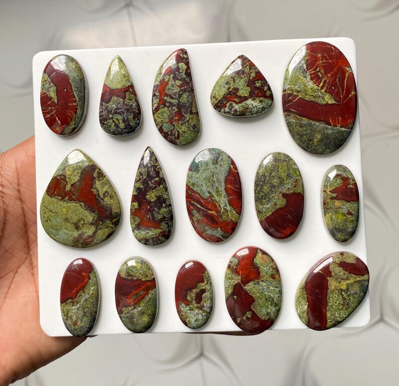 May include: A collection of fourteen polished cabochons arranged on a white rectangular display board. Each stone features a unique pattern of deep red and olive green swirls, with smooth, rounded edges in various oval, teardrop, and irregular shapes.
