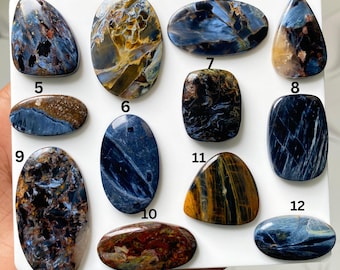 May include: A collection of twelve polished gemstones arranged on a white surface, numbered 1 through 12. The stones feature varied patterns and colors including deep blues, golden browns, blacks, and rust tones, with smooth, rounded edges.