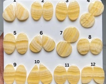 Può includere: Una collezione di dodici paia di cabochon in calcite gialla a bande lucidata disposti su un pannello bianco. Ogni pietra presenta strisce orizzontali color crema e giallo. Le coppie sono numerate da 1 a 12 e mostrano varie forme, tra cui gocce, ovali e cerchi con superfici lisce e lucide.