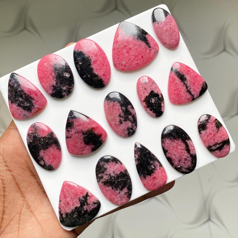 May include: A collection of fourteen polished rhodonite cabochons arranged on a white display board. Each stone features a distinct pattern of vibrant pink and deep black mineral inclusions, with various shapes including ovals, triangles, and teardrops.