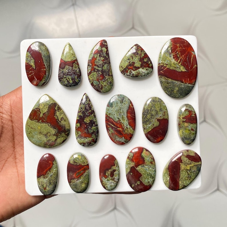 May include: A collection of fourteen polished cabochons arranged on a white rectangular display board. Each stone features a distinct pattern of deep red and moss green mineral inclusions, with various shapes including ovals, teardrops, and rounded forms.
